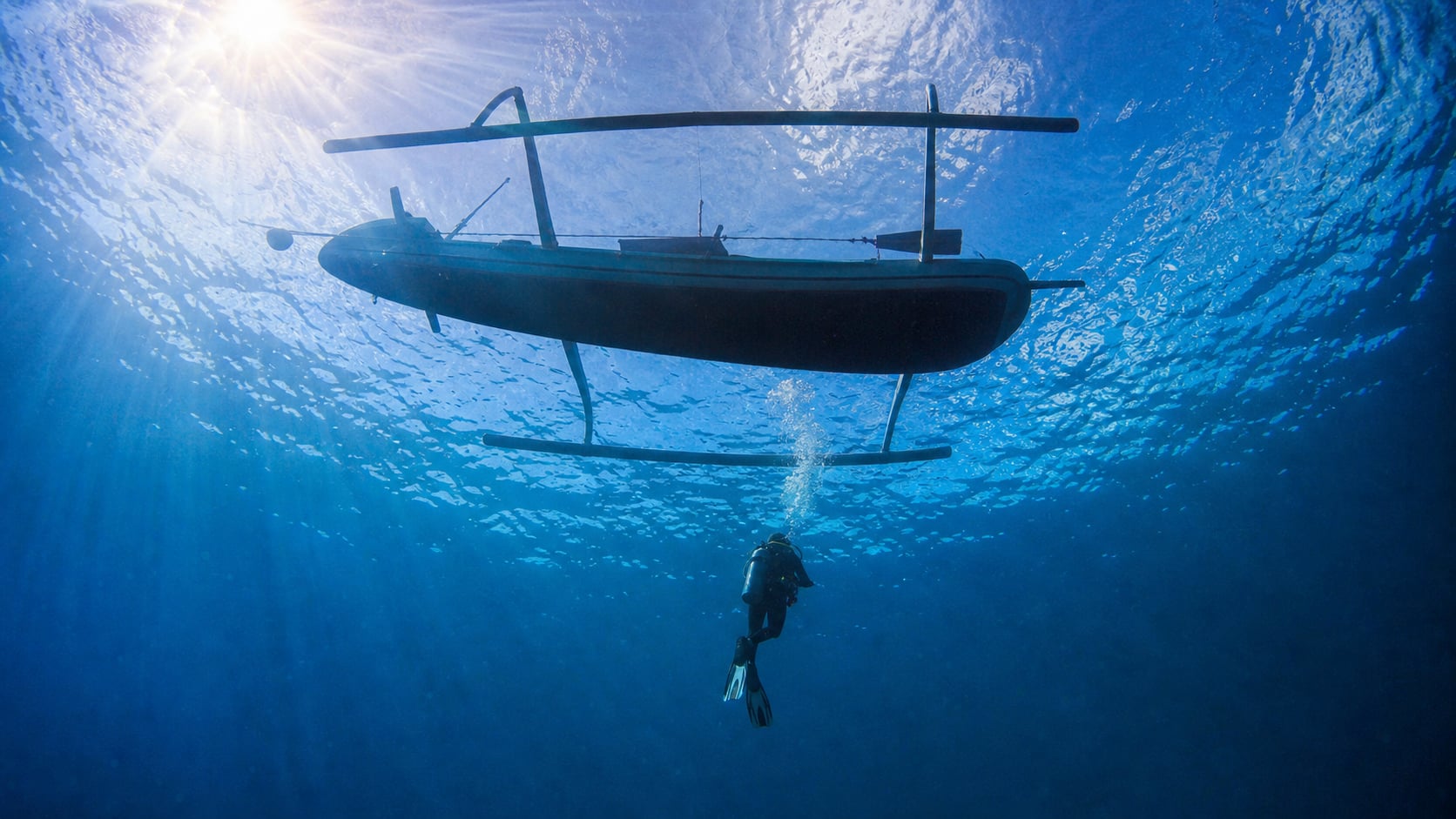 Balinesisches Auslegerboot von unten gegen die Wasseroberfläche, darunter ein abtauchender Taucher